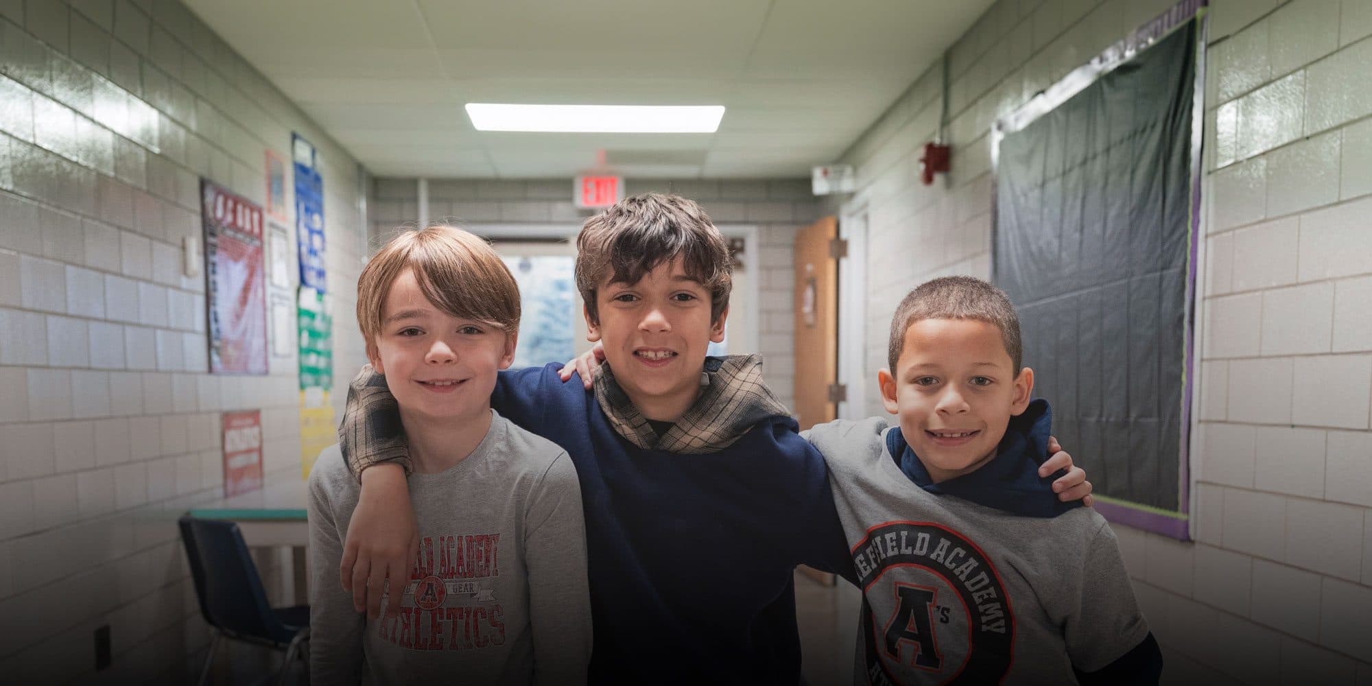 Three smiling boys with arms around each other in a school hallway.